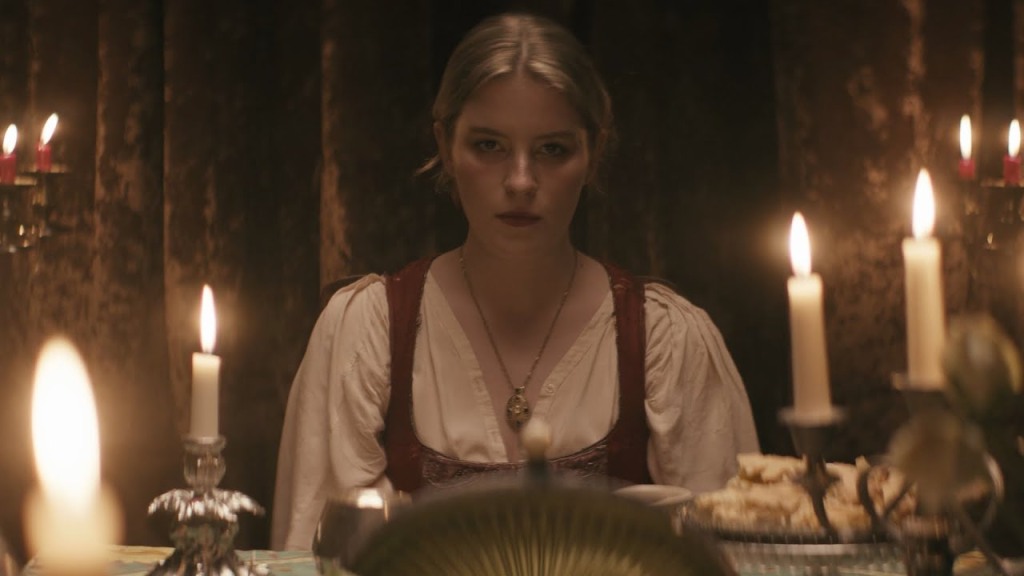 A white woman with blonde hair and blue eyes sits in a candlelit kitchen, wearing a peasant blouse.